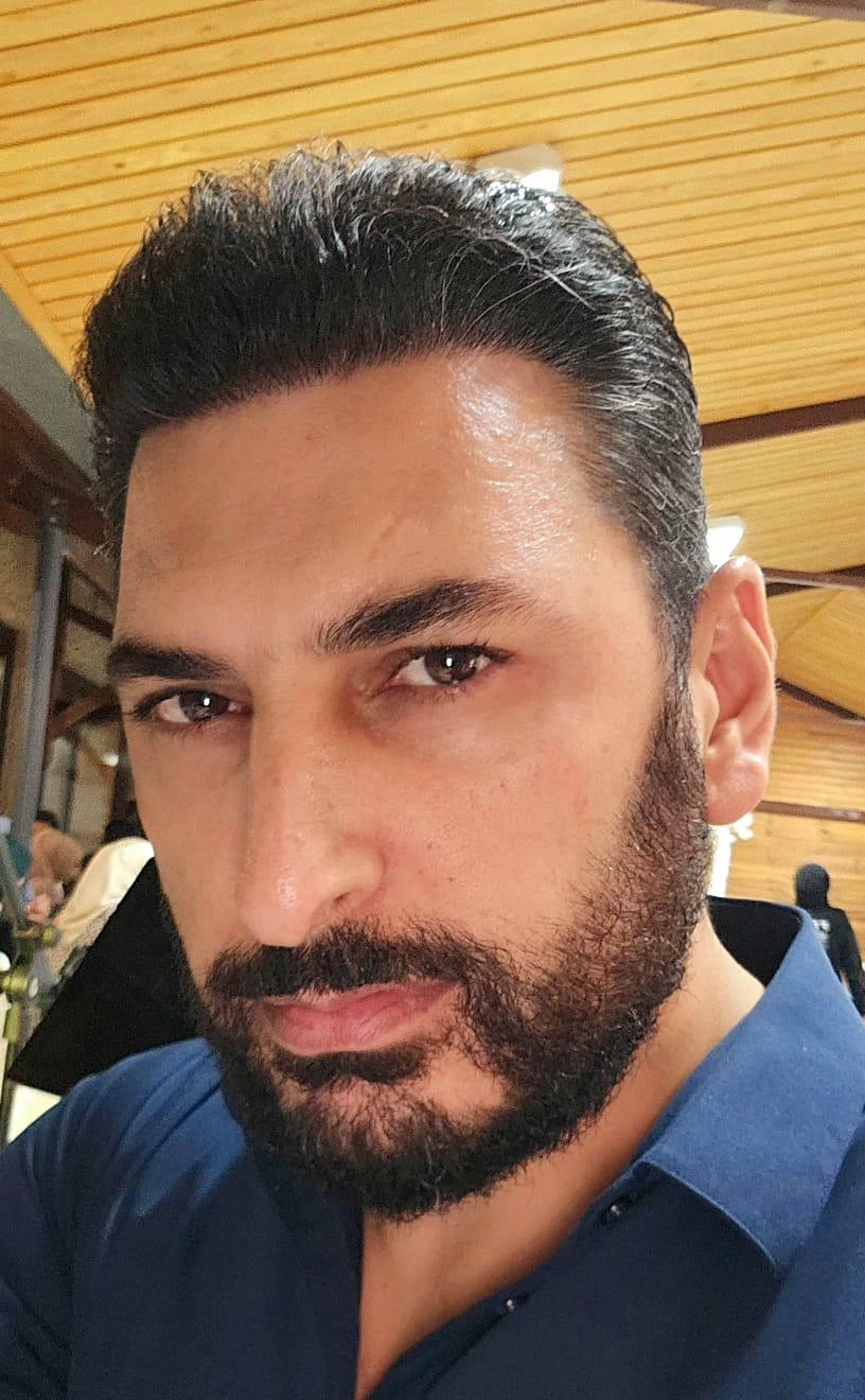 Ein Mann mit dunklem Haar und Bart schaut ernst in die Kamera, trägt ein blaues Hemd