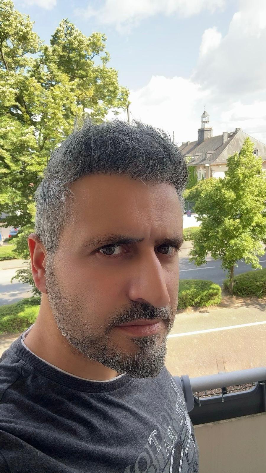 Ein Mann mit graumelierten Haaren schaut ernst, im Hintergrund Bäume und Gebäude