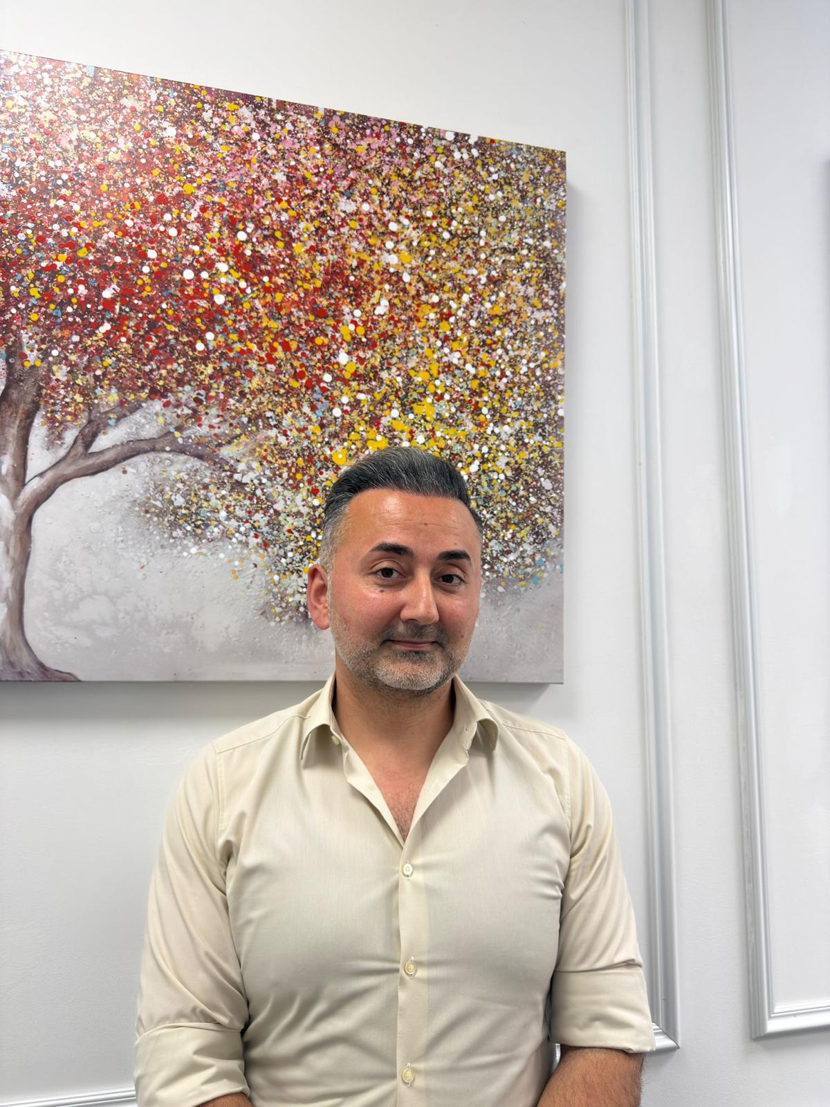 Ein Mann mit zurückgekämmten dunklen Haaren und Bart steht lächelnd vor buntem Baumgemälde