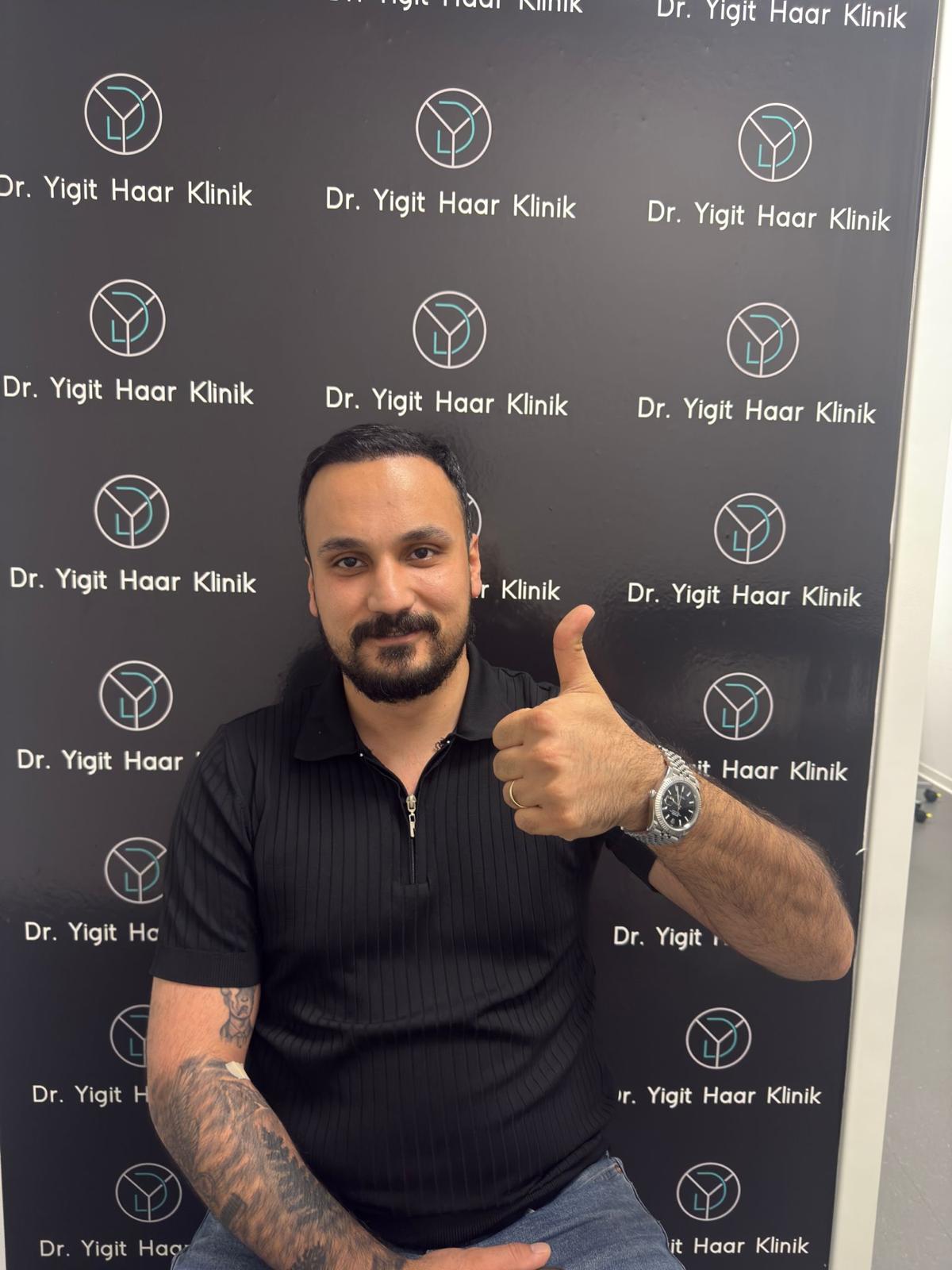 Mann mit dunklen Haaren und Bart lächelt und zeigt Daumen hoch vor schwarzer Wand