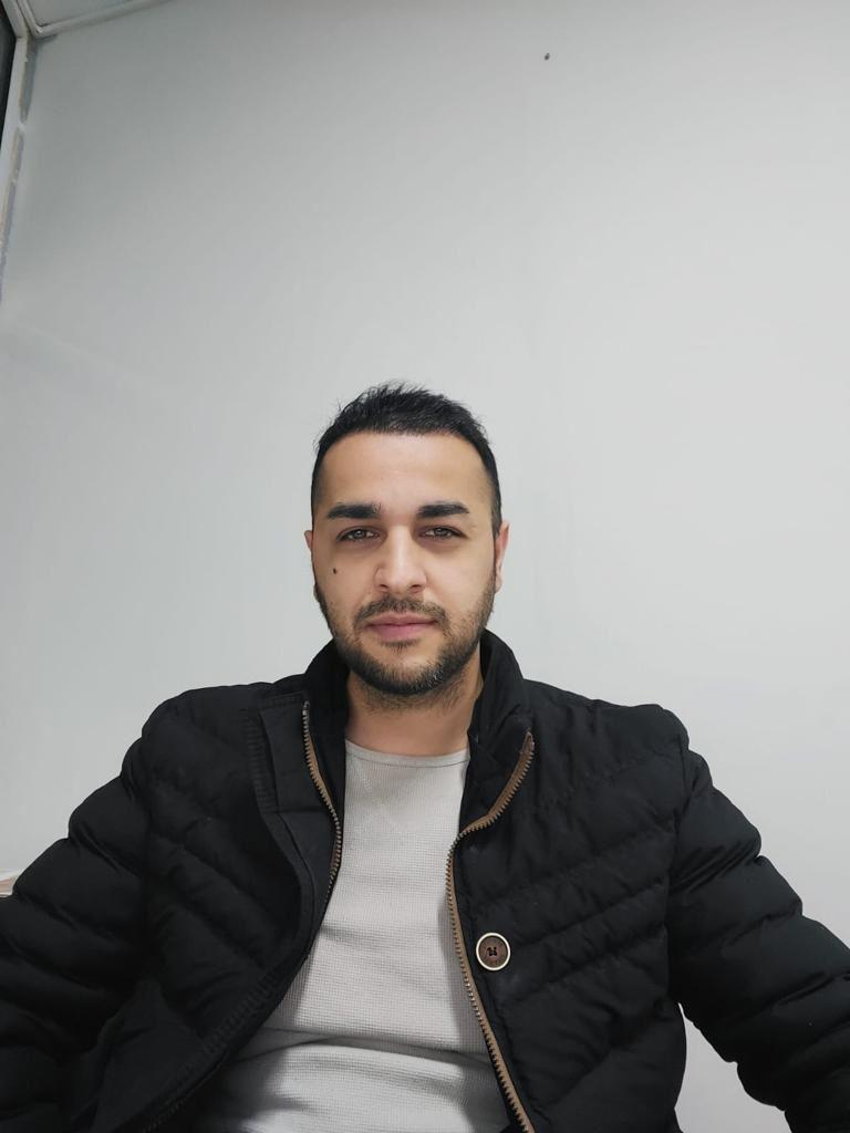 Ein junger Mann mit dunklem Haar und Bart blickt ruhig in die Kamera, trägt eine schwarze Jacke