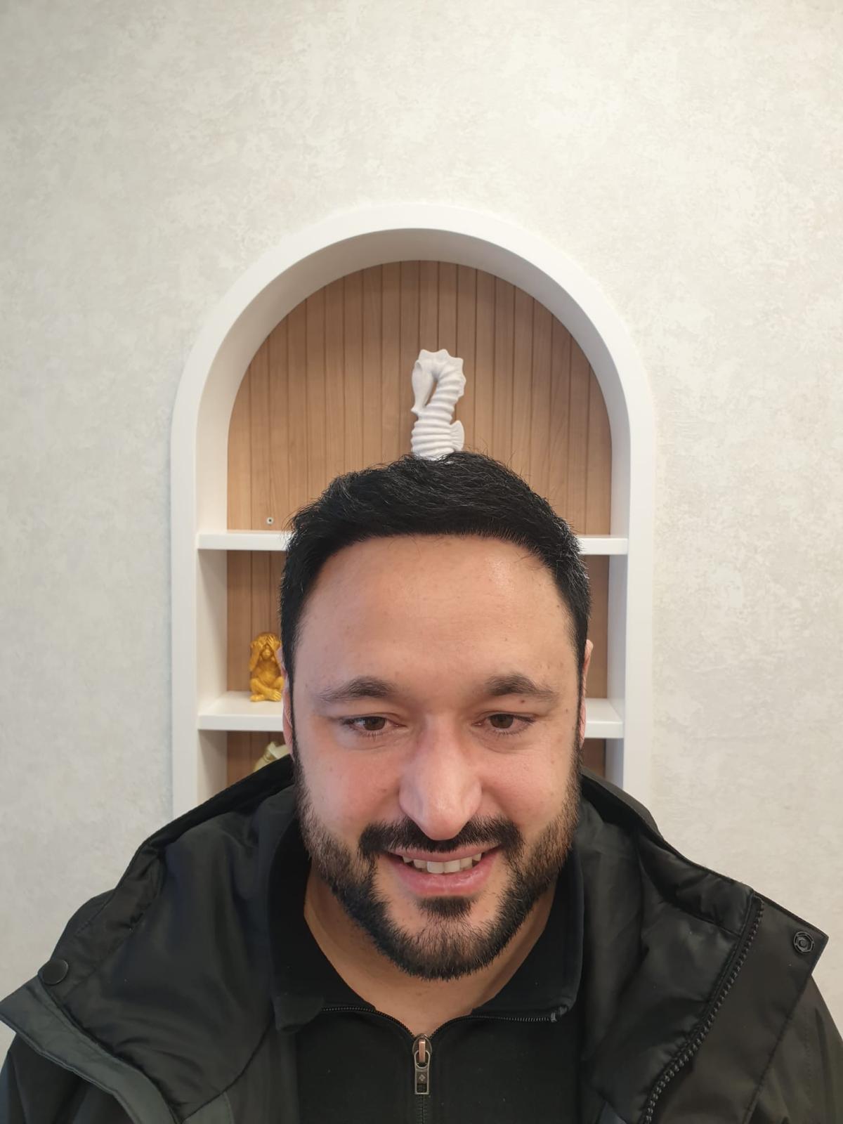 Ein lächelnder Mann mit dunklem Haar und Bart vor einem hellen Regal mit Skulpturen