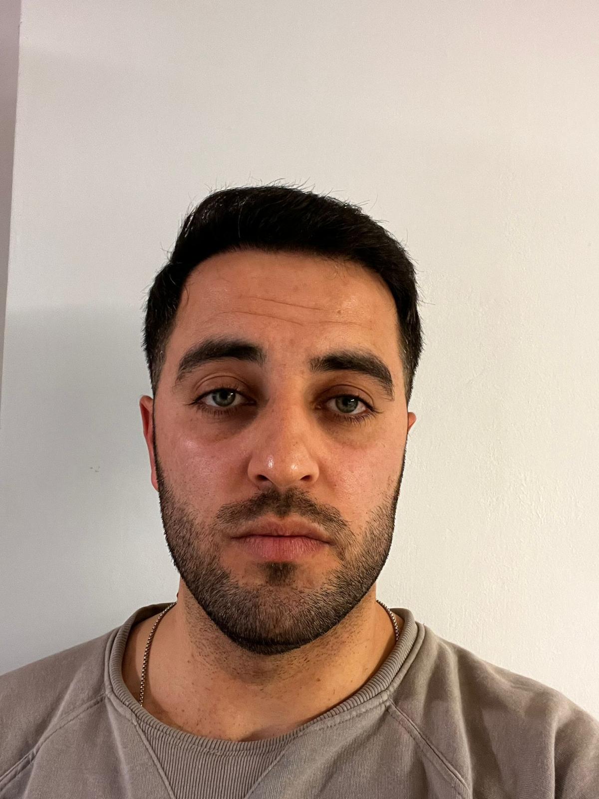 Junger Mann mit dunklen Haaren blickt neutral in die Kamera vor heller Wand