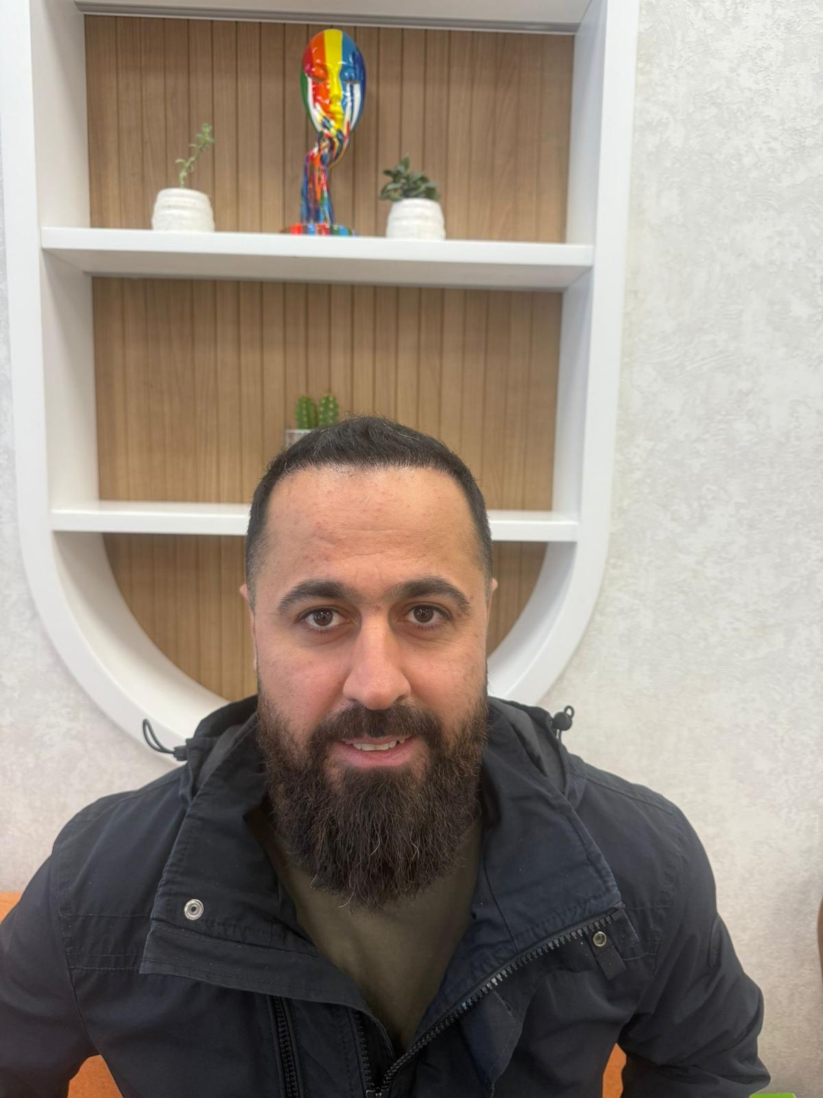 Ein Mann mit dunklem Bart lächelt leicht vor einem Regal mit bunter Skulptur