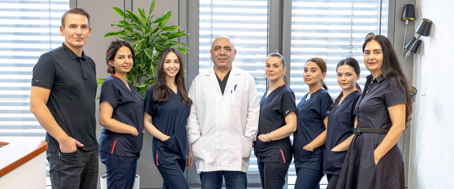 Sieben Personen stehen lächelnd nebeneinander, teils in dunkler Kleidung, teils im weißen Kittel